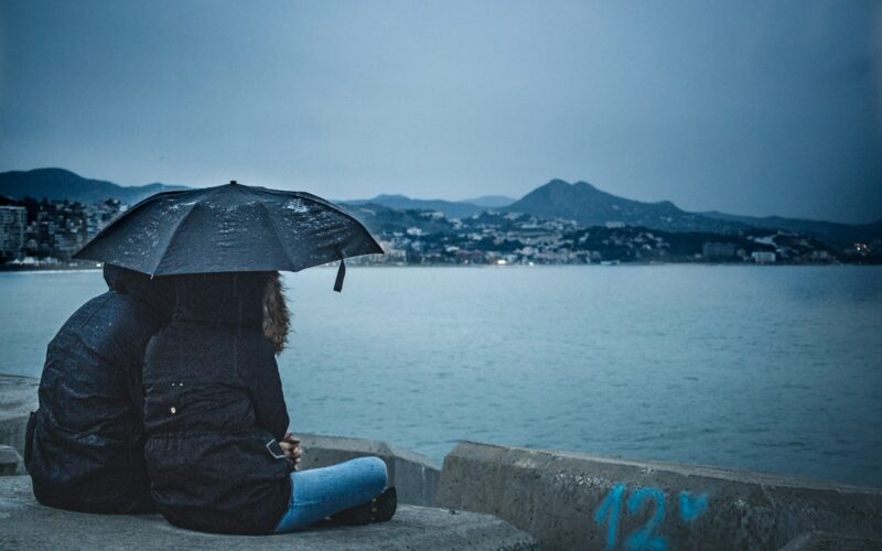 couple holding black umbrella while sitting beside calm water